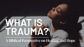 A woman sits with her knees pulled close, looking upward with a pained expression as dramatic lighting casts shadows around her. Text on the image reads: ‘What Is Trauma?’ and ‘A Biblical Perspective on Healing and Hope.’