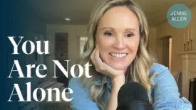 A woman smiles warmly while sitting at a desk with a microphone in front of her, resting her chin on her hand. Text on the image reads: ‘You Are Not Alone.’ The background shows a cozy living room setting.